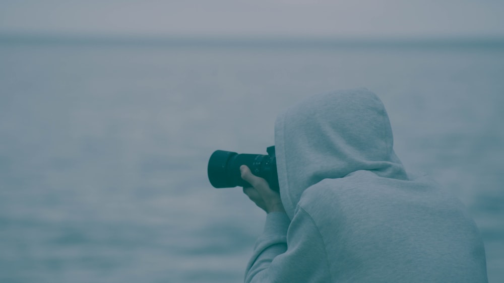person wearing hoodie holding camera taking photo near body of water during daytime