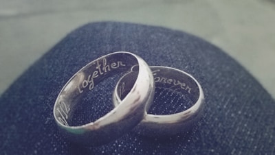 Two silver rings engraved with the words 'Together' and 'Forever' rest on a dark, textured surface, possibly denim. The rings are intertwined, signifying connection and unity.