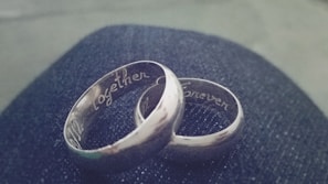 Two silver rings engraved with the words 'Together' and 'Forever' rest on a dark, textured surface, possibly denim. The rings are intertwined, signifying connection and unity.