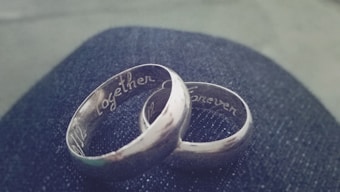 Two silver rings engraved with the words 'Together' and 'Forever' rest on a dark, textured surface, possibly denim. The rings are intertwined, signifying connection and unity.
