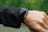 A person is wearing a smartwatch on their wrist. The smartwatch has a black strap and a rectangular face, and it appears to be turned off or in sleep mode. The person is wearing a long-sleeved black jacket, and the background is a soft-focus green, suggesting an outdoor setting.