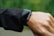 A person is wearing a smartwatch on their wrist. The smartwatch has a black strap and a rectangular face, and it appears to be turned off or in sleep mode. The person is wearing a long-sleeved black jacket, and the background is a soft-focus green, suggesting an outdoor setting.