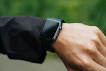 A person is wearing a smartwatch on their wrist. The smartwatch has a black strap and a rectangular face, and it appears to be turned off or in sleep mode. The person is wearing a long-sleeved black jacket, and the background is a soft-focus green, suggesting an outdoor setting.