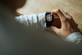 A person interacting with a smartwatch on their wrist, tapping the screen where multiple colorful app icons are visible. The individual is wearing a long-sleeved shirt, and the setting appears to be a wooden table.