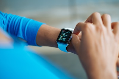 A person interacts with a smartwatch on their wrist, displaying a colorful screen featuring app icons and text. The watch is secured with a white strap, and the individual's attire includes a blue long-sleeved shirt. The background is softly blurred, focusing attention on the watch and the hand.