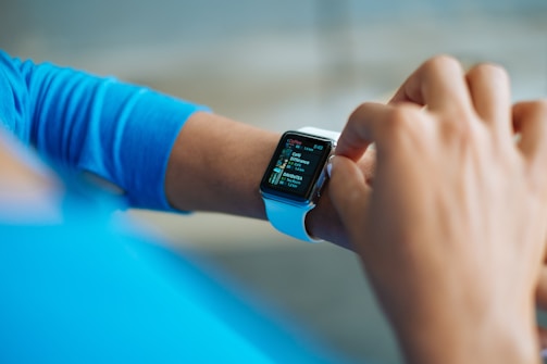 A person interacts with a smartwatch on their wrist, displaying a colorful screen featuring app icons and text. The watch is secured with a white strap, and the individual's attire includes a blue long-sleeved shirt. The background is softly blurred, focusing attention on the watch and the hand.