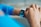 A person interacts with a smartwatch on their wrist, displaying a colorful screen featuring app icons and text. The watch is secured with a white strap, and the individual's attire includes a blue long-sleeved shirt. The background is softly blurred, focusing attention on the watch and the hand.