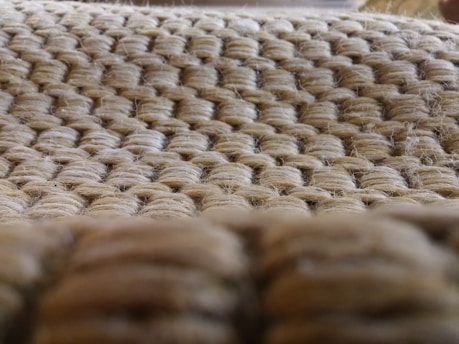 A close-up view of a woven texture made from light brown, natural fibers. The pattern is tightly knit, with a repeating sequence of interlaced strands creating a uniform appearance.