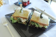 Two sandwiches with golden toasted bread, layered with green lettuce and other fillings, are presented on a square black plate. Each sandwich is garnished with a pickle skewered on top. The sandwiches are accompanied by a shiny silver fork and knife on a white tablecloth. In the background, there are small jars of condiments and a portion of fries.