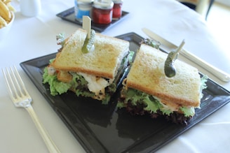 Two sandwiches with golden toasted bread, layered with green lettuce and other fillings, are presented on a square black plate. Each sandwich is garnished with a pickle skewered on top. The sandwiches are accompanied by a shiny silver fork and knife on a white tablecloth. In the background, there are small jars of condiments and a portion of fries.