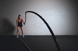 A person is engaged in an intense workout, using battle ropes in a dimly lit gym. The individual is wearing a black athletic outfit and blue sneakers, standing against a plain white brick wall. The ropes are in motion, creating a dynamic effect.