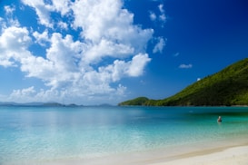 A serene beach scene with turquoise waters gently lapping against a white sand shore. Lush green hills rise in the background under a vibrant blue sky dotted with fluffy white clouds. One person is seen wading into the water, adding a sense of tranquility to the setting.