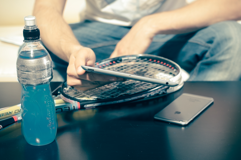 person sitting while using iPhone 8 and tennis racket beside energy drink bottle