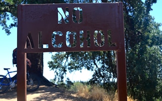 A metal sign with the words 'NO ALCOHOL' cut out stands in the foreground. Behind it, a cyclist with a blue bicycle is visible under the shade of large trees. Sunlight filters through the leaves, creating a dappled light effect on the ground.
