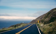 A scenic coastal road curves along a hillside with the ocean visible to the left. The sky is somewhat overcast, with clouds hovering above the distant horizon. A road sign indicates a slower speed, set against the lush green vegetation on the hillside.
