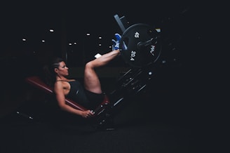 A person is exercising on a leg press machine in a gym setting. The environment is dimly lit, highlighting the subject's concentration and effort. The person is wearing a tank top and shorts, with one foot squared against the footplate, pushing weights labeled 45 and 20.4.