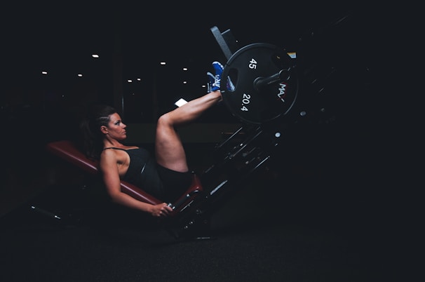 A person is exercising on a leg press machine in a gym setting. The environment is dimly lit, highlighting the subject's concentration and effort. The person is wearing a tank top and shorts, with one foot squared against the footplate, pushing weights labeled 45 and 20.4.