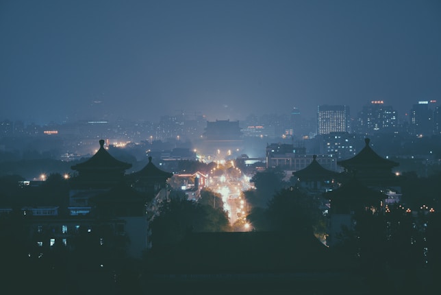 A cityscape at night with illuminated buildings and traditional pagoda-style rooftops in the foreground. The scene is enveloped in a bluish haze, with city lights creating a warm glow against the cool surroundings.