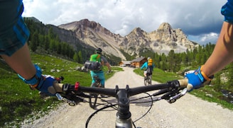 A cyclist's view of riding down a gravel path flanked by lush green grass and rocky terrain. Ahead, two additional cyclists are pedaling towards a cabin nestled amidst tall, pointed mountains with scattered trees. The sky is partly cloudy, casting a mix of shadows and sunlight over the scene.
