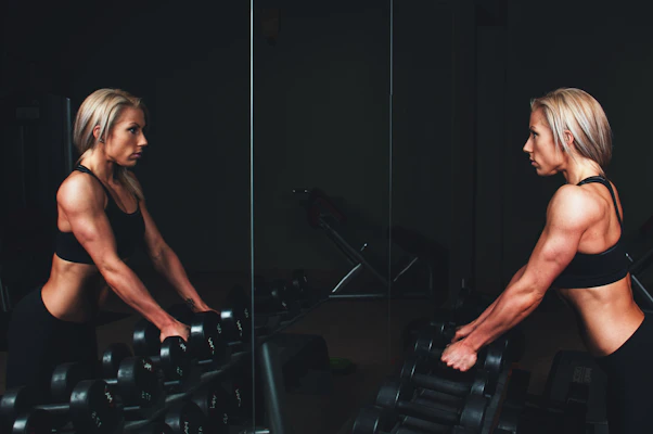 woman wearing black top top holding black dumbbells standing in front of mirror