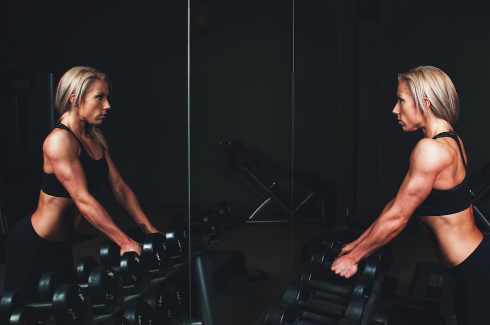 woman wearing black top top holding black dumbbells standing in front of mirror