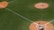 A baseball game in progress on a well-maintained field. A pitcher is delivering a pitch from the mound toward the batter at home plate. The batter is in position to swing, while the umpire and catcher are in place behind him. The scene is captured from an aerial view, clearly displaying the diamond layout on a sunny day.