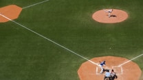 A baseball game in progress on a well-maintained field. A pitcher is delivering a pitch from the mound toward the batter at home plate. The batter is in position to swing, while the umpire and catcher are in place behind him. The scene is captured from an aerial view, clearly displaying the diamond layout on a sunny day.