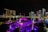 A nighttime view of Marina Bay in Singapore, featuring the iconic Marina Bay Sands hotel with its three towers and boat-shaped roof. Vibrant lights from surrounding skyscrapers reflect off the water, creating a dazzling cityscape. In the foreground, a structure with a purple canopy is illuminated, adding a vibrant accent to the scene.