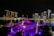 A nighttime view of Marina Bay in Singapore, featuring the iconic Marina Bay Sands hotel with its three towers and boat-shaped roof. Vibrant lights from surrounding skyscrapers reflect off the water, creating a dazzling cityscape. In the foreground, a structure with a purple canopy is illuminated, adding a vibrant accent to the scene.