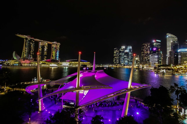 A nighttime view of Marina Bay in Singapore, featuring the iconic Marina Bay Sands hotel with its three towers and boat-shaped roof. Vibrant lights from surrounding skyscrapers reflect off the water, creating a dazzling cityscape. In the foreground, a structure with a purple canopy is illuminated, adding a vibrant accent to the scene.