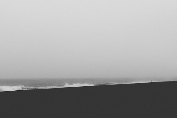 A minimalist black and white photograph capturing a solitary person standing on a vast beach, with waves crashing in the background and a foggy sky.