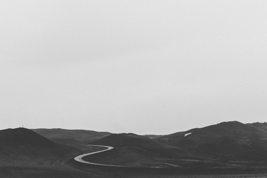 A black and white landscape featuring a winding road cutting through a series of dark hills. The sky is overcast with a smooth, gray tone, creating a calm and minimalistic scene.