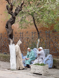 Three people are interacting in an outdoor setting with a rustic background. A person in a striped robe leans against a partially painted tree, while two others sit on a blue and white ornate bench, engaged in conversation. The area is enclosed by intricate wrought iron fencing, and a planter with flowers is placed nearby. A tree with fresh green leaves provides shade, and the walls behind them are earthy-toned, suggesting an older architectural style.