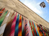 A series of colorful fabric rolls hanging vertically against an aged, rustic wall with a clear blue sky above. The fabrics display a variety of vibrant patterns and colors, adding a lively contrast to the muted tones of the weathered wall. A lamppost is visible on the upper right corner, contributing to the urban setting.