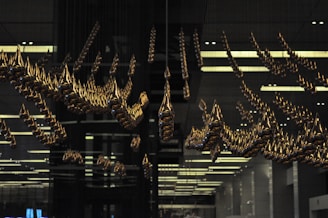An intricate installation of golden teardrop-shaped elements suspended from the ceiling in a dark, modern interior space, reflecting light and casting unique shadows. The space has a structured, symmetrical design, contributing to a sense of order and precision.