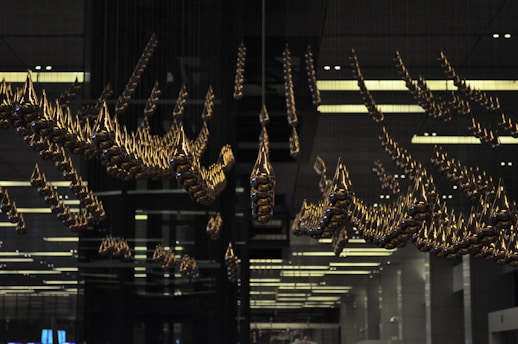 An intricate installation of golden teardrop-shaped elements suspended from the ceiling in a dark, modern interior space, reflecting light and casting unique shadows. The space has a structured, symmetrical design, contributing to a sense of order and precision.