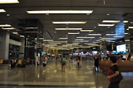 A spacious airport terminal with high ceilings and modern lighting. Several people are walking or sitting, with some standing in line at a counter. The area is adorned with digital screens and signage. Luggage and travel bags are visible, and the floor is reflective, adding a sense of openness to the space.