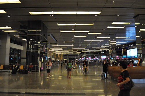 A spacious airport terminal with high ceilings and modern lighting. Several people are walking or sitting, with some standing in line at a counter. The area is adorned with digital screens and signage. Luggage and travel bags are visible, and the floor is reflective, adding a sense of openness to the space.