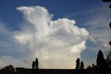 A large, billowing white cloud dominates the sky, with patches of blue visible around it. The cloud appears thick and fluffy, creating a dramatic and almost surreal effect. Silhouettes of tall, thin trees are visible in the foreground, along with the top of a building.