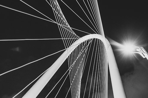 A striking architectural structure featuring cables and arches, illuminated dramatically against a dark sky. The cables radiate outward from the top, creating an intricate pattern.