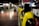 A row of yellow public telephones is lined along a dimly lit corridor, with a person in the background using one of the phones. The environment suggests an urban setting, possibly a subway or train station. The lights create a reflection on the polished floor.