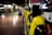 A row of yellow public telephones is lined along a dimly lit corridor, with a person in the background using one of the phones. The environment suggests an urban setting, possibly a subway or train station. The lights create a reflection on the polished floor.