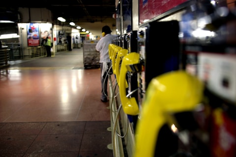 A row of yellow public telephones is lined along a dimly lit corridor, with a person in the background using one of the phones. The environment suggests an urban setting, possibly a subway or train station. The lights create a reflection on the polished floor.