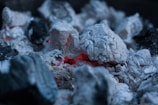 Hot charcoal embers are surrounded by white ash, with a few glowing red areas indicating residual heat. The texture of the charcoal and ash is coarse and uneven, with various lumps and fragments visible.