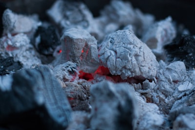 Hot charcoal embers are surrounded by white ash, with a few glowing red areas indicating residual heat. The texture of the charcoal and ash is coarse and uneven, with various lumps and fragments visible.