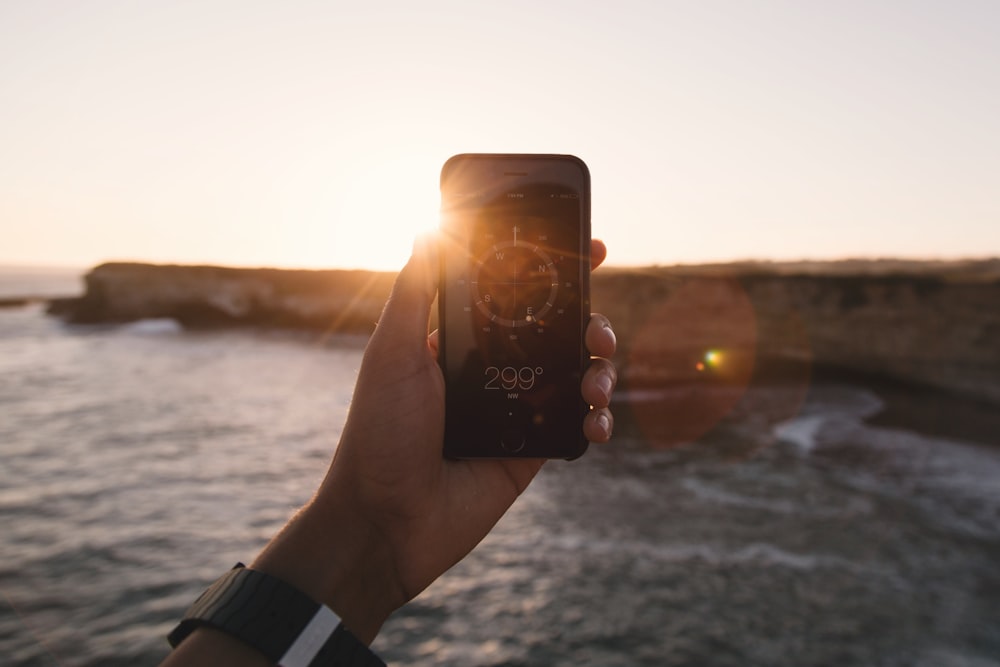 person holding black phone beside body of water under white sky during golden hour