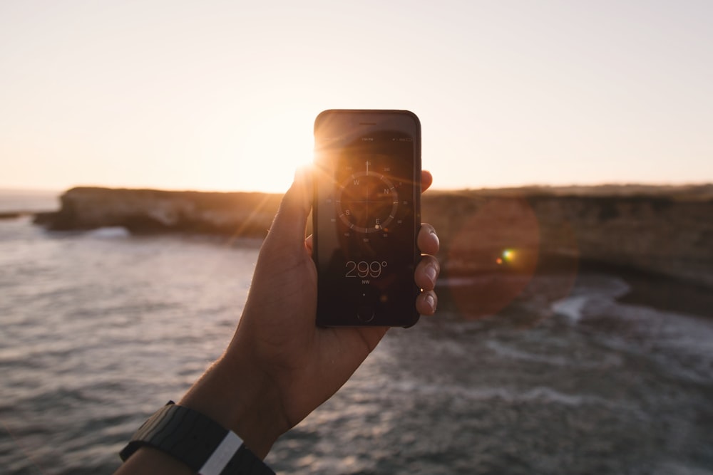 Person mit schwarzem Telefon neben Gewässer unter weißem Himmel während der goldenen Stunde