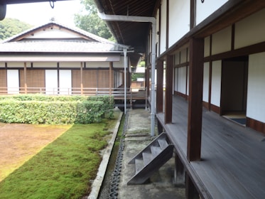 A traditional Japanese wooden house featuring an exterior hallway with wooden floors and sliding doors. The building is surrounded by a meticulously maintained garden with green hedges and a carpet of moss. The roof has a classic Japanese architecture style with gray tiles.
