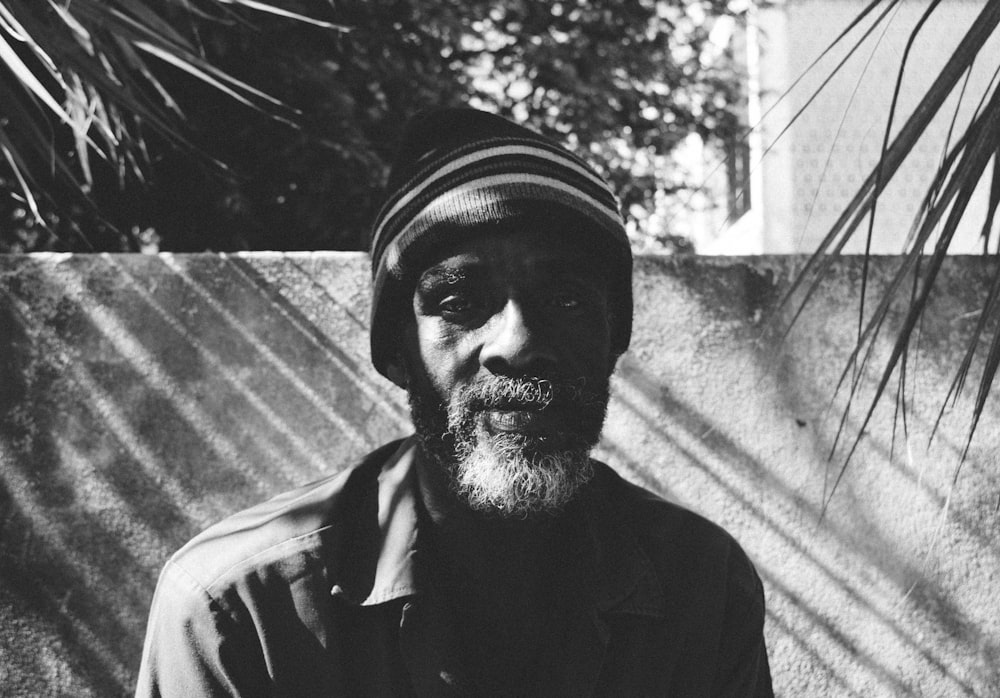 grayscale photo of man wearing beanie in front of concrete wall and between two linear-leafed plants