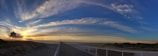 A panoramic view of a boardwalk leading to an expansive beach, with a wooden hut on stilts to the left and a vast sky painted with dramatic, sweeping clouds during sunset.